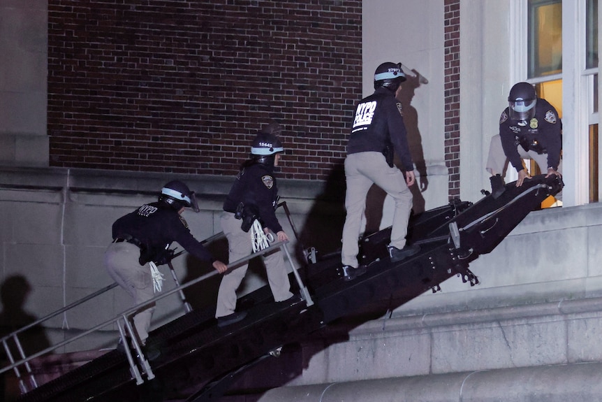 Three riot police climb up a ramp to a building, a fourth police officer holds it down