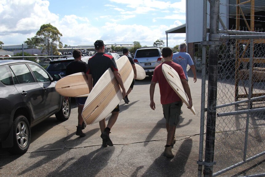 Five men walking past a chain link fence into a workshop with wooden surfboards tucked under their arms.