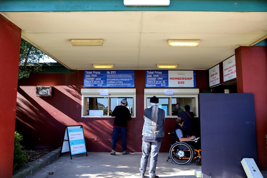 Two men standing and one man in a wheelchair outside a booking window of a racecourse that has signs for COVID-19 vaccinations 