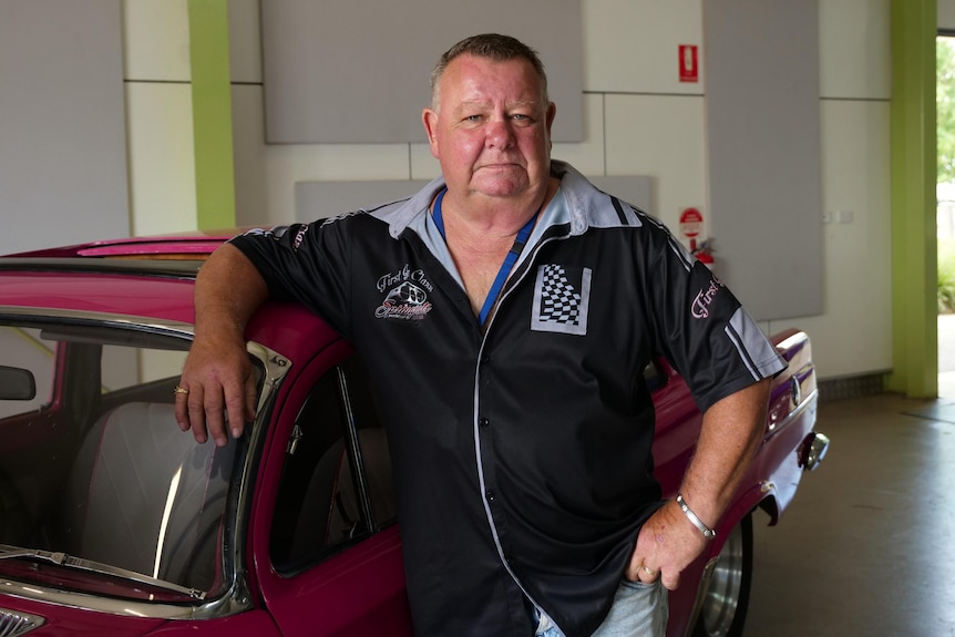 A middle-aged heavyset Caucasian man leans on a red car in a garage, wears a black and grey tee. Not smiling.