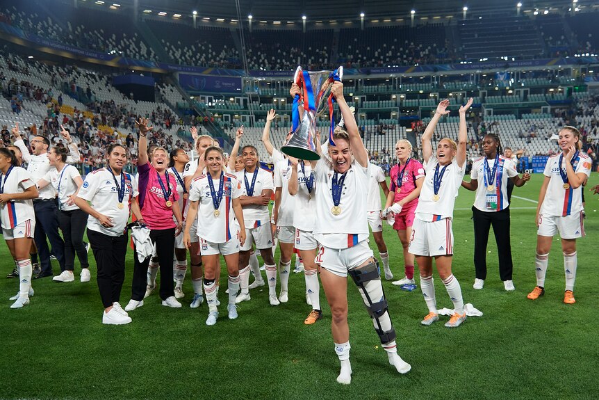 Australian footballer Ellie Carpenter lifts a cup above her head while wearing a leg brace as her teammates cheer.