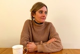 Katie wears a brown jumper, sitting at a kitchen table with a white cup of tea, looking pensively away from the camera.