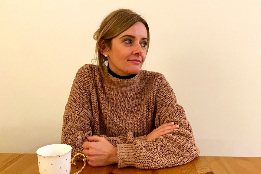 Katie wears a brown jumper, sitting at a kitchen table with a white cup of tea, looking pensively away from the camera.