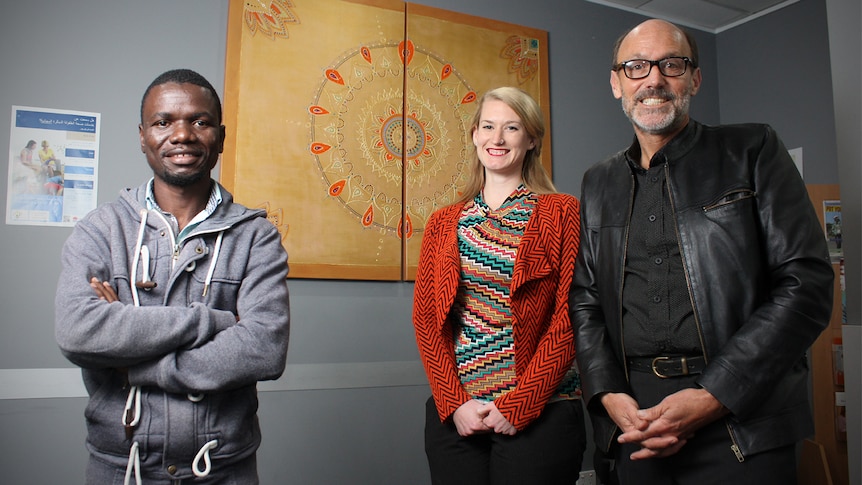 Jay Jay Ganwaye, Monique Bolus and Rob Goodfellow stand in front of cultural artwork smiling at the camera.