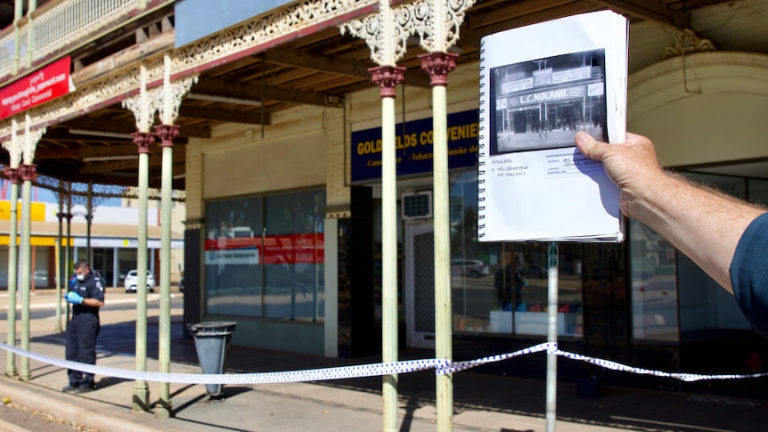 A hand holding a notebook with an old picture in front of the building photographed, with police tape around it 