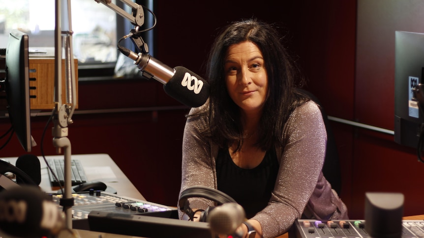 Tara is in a radio studio, leaning forward on the desk next to a microphone. She has dark hair and a kind look on her face.