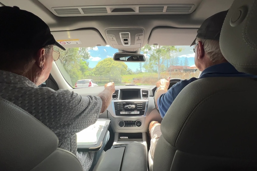 View from rear seat of a car of two men driving, the passenger pointing with a map on his lap.