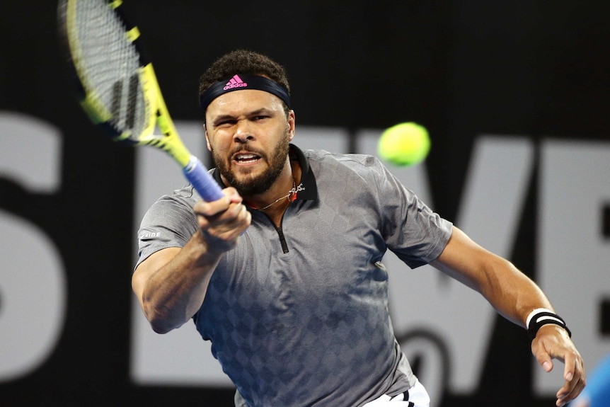 Jo-Wilfried Tsonga hits a forehand while wearing a grey shirt and a black headband with a grimace expression.