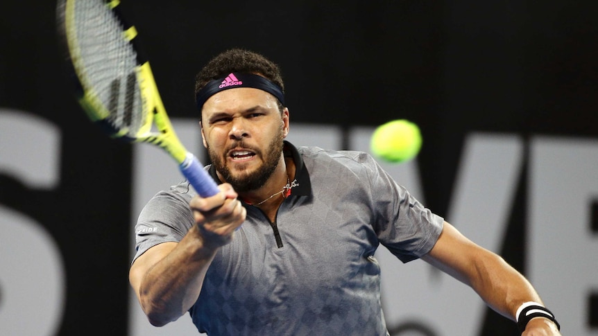 Jo-Wilfried Tsonga hits a forehand while wearing a grey shirt and a black headband with a grimace expression.