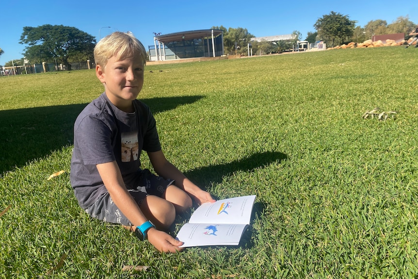 Smiling blond boy sitting in park, book on grass in front, wears purple tee, blue strap watch, looks at camera.