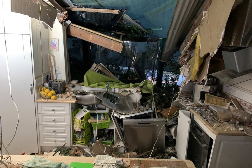 A kitchen is destroyed and a wall is caved in and covered by a tarp.