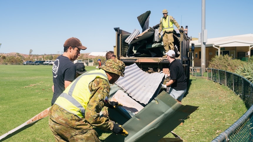 A man wearing army fatigues in a truck as people load pieces of corrugated fencing up.