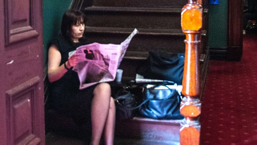 A female reporter sitting on the bottom step of a wooden staircase in a courtroom, reading a newspaper.