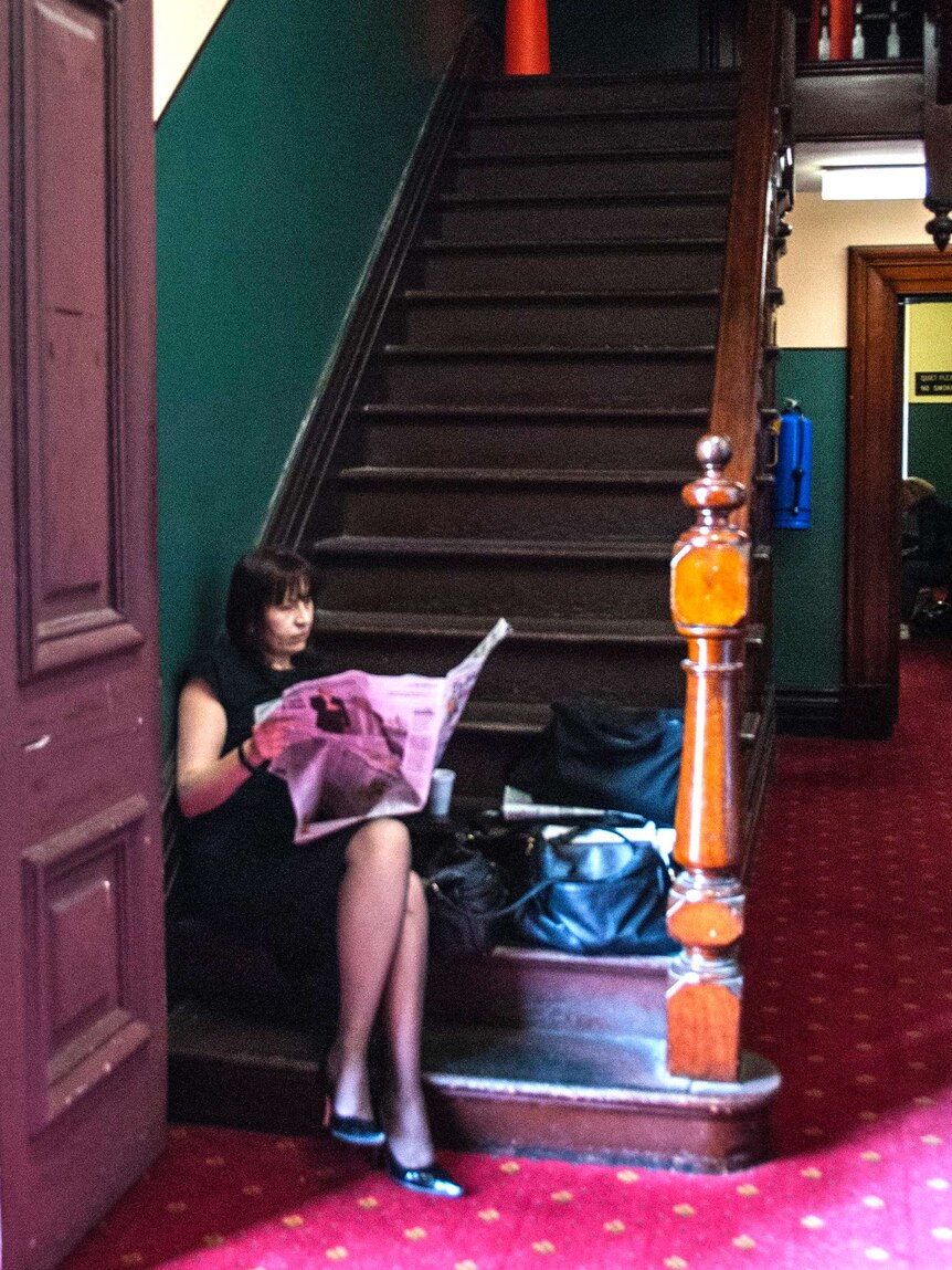 A female reporter sitting on the bottom step of a wooden staircase in a courtroom, reading a newspaper.