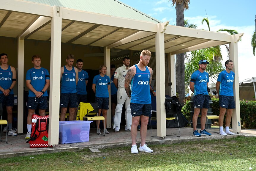 England cricket players, led by Ben Stokes, bow their heads as they stand under an awning before a Test.