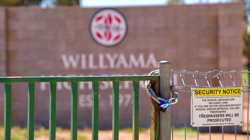 A lock wrapped around a green gate next to a security notice, behind red brick wall with Williyama High School with logo.