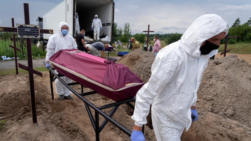 Men in Hazmat suits carry a covered coffin away from a truck past a row of open graves