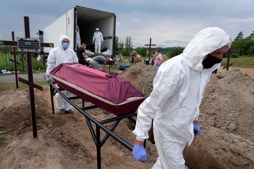 Men in Hazmat suits carry a covered coffin away from a truck past a row of open graves