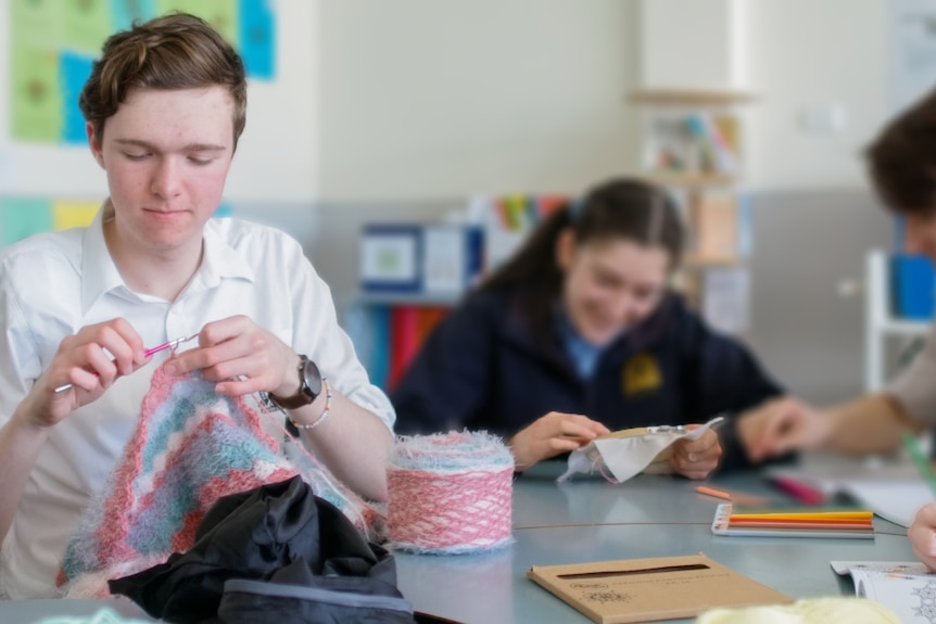 Jorden crochets a blanket while sat in a classroom with three other people seen doing their own crafts too.