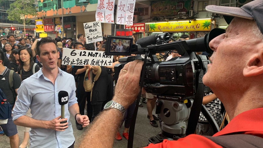 Birtles holding microphone looking and talking to camera with crowd of protestors behind him.