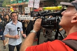 Birtles holding microphone looking and talking to camera with crowd of protestors behind him.