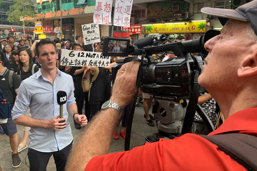 Birtles holding microphone looking and talking to camera with crowd of protestors behind him.