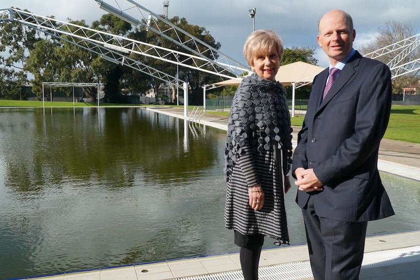 A blonde woman in a grey shawl and a man in a suit stand in front of an outdoor pool