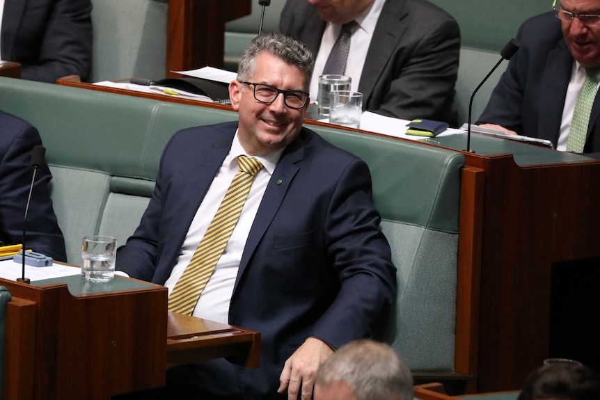 A grey-haired man sitting in Parliament, smiling, wearing a blue suit and gold stripy tie.