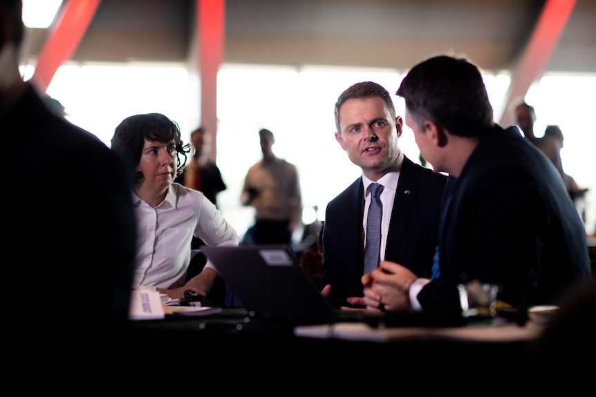 A woman and two men sit at a desk talking. Behind them are people standing, out of focus