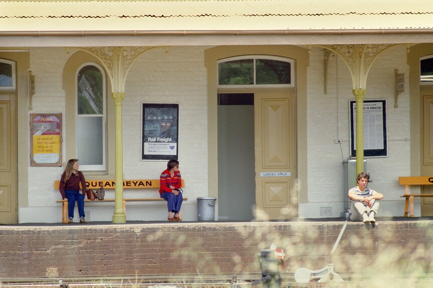 Three people sitt waiting at a regional train station.