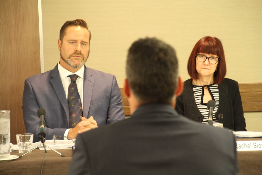 A man and woman in suits sitting at table with man's back to them.