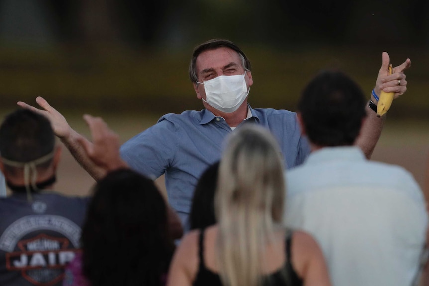 A smiling man wearing a face mask raises his hands while holding a banana and talking to a crowd.