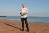 A photo of Michael Wells standing on the shore of Cullen Bay Beach, holding a photo of his late sister.
