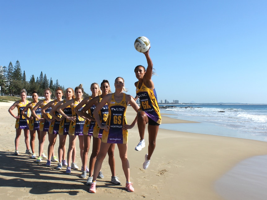 A line of netball players at the beach with one jumping with a netball at the front.