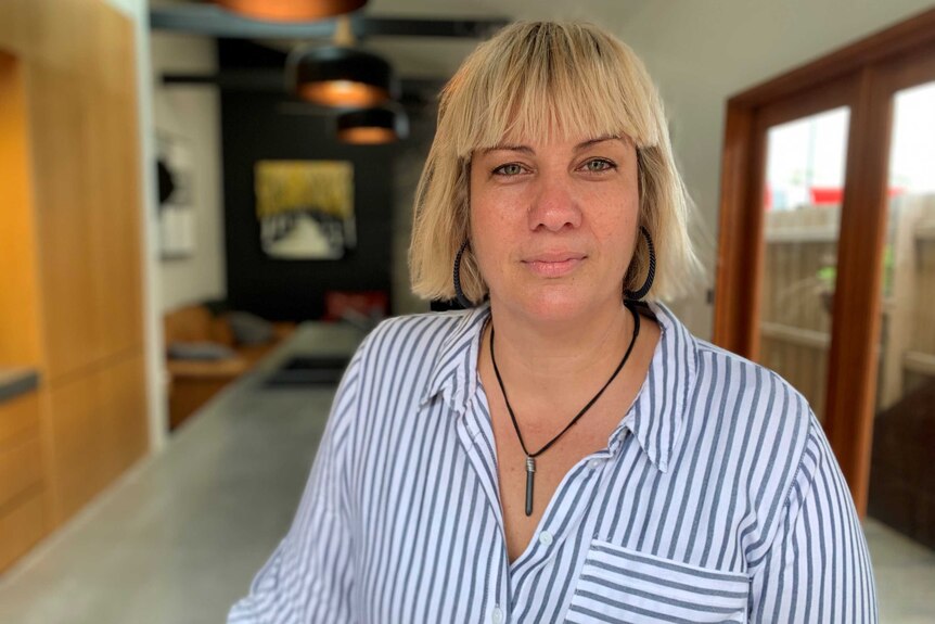 A young woman in an open-necked grey-striped shirt stands in the kitchen of a townhouse looking unsmiling at the camera.