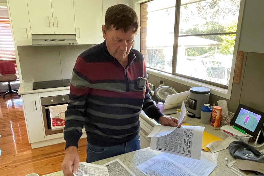 Man at a kitchen bench looks over photocopies of newspapers
