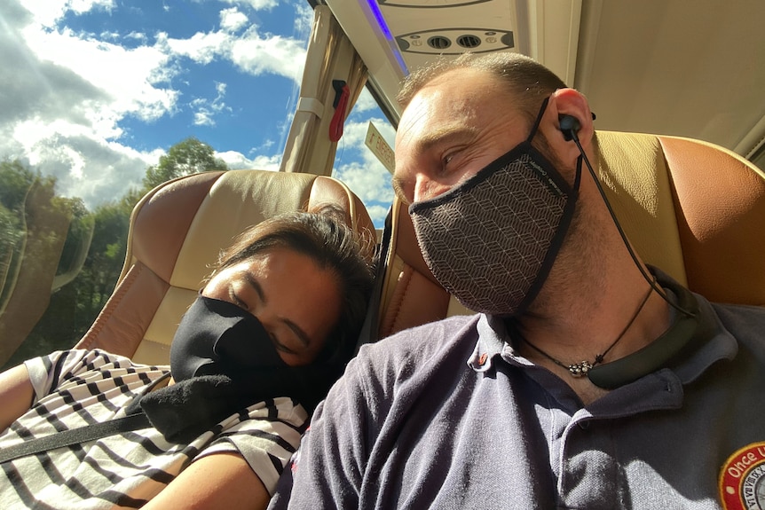 A woman wearing a mask sleeping on the shoulder of a masked man on a bus