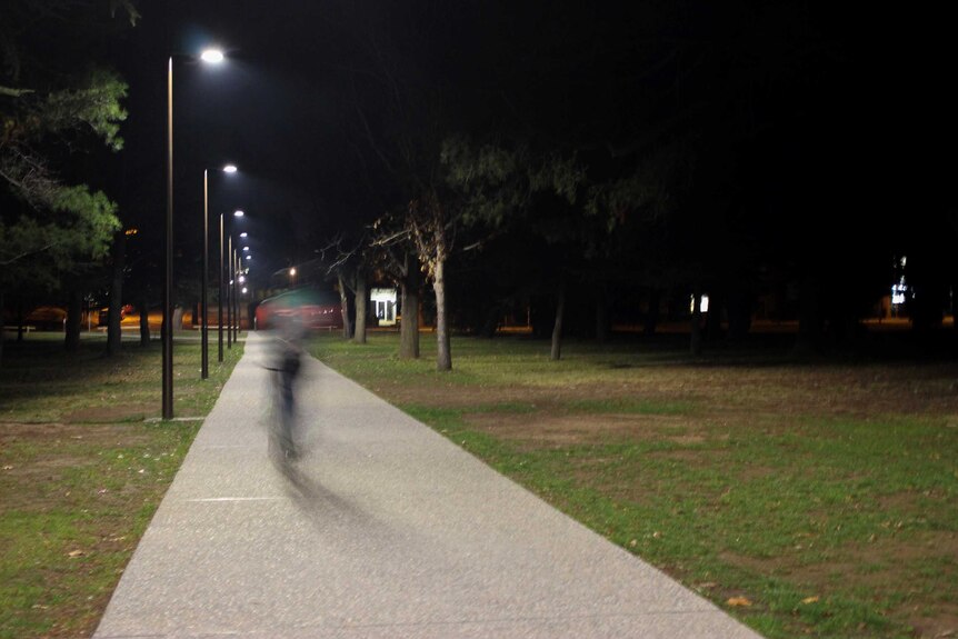 A cyclist, unidentifiable through motion blur, rides on a lit path through a Canberra park.