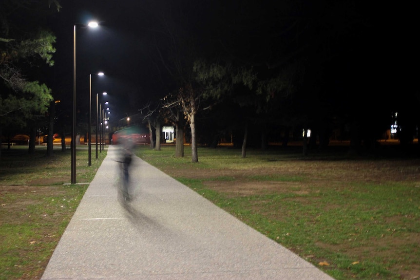 A cyclist, unidentifiable through motion blur, rides on a lit path through a Canberra park.