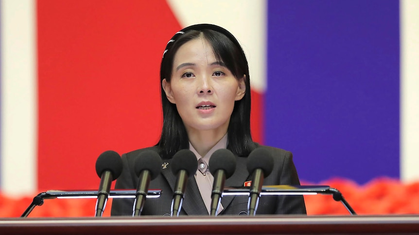 Kim Yo Jong speaking into a lecturn with 5 microphones, wearing a black blazer. 