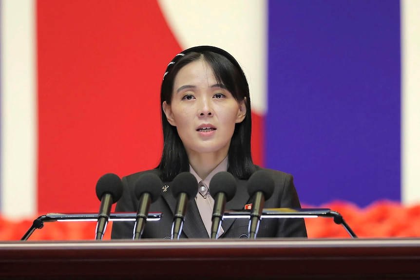 Kim Yo Jong speaking into a lecturn with 5 microphones, wearing a black blazer. 