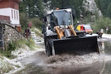 A worker uses a digger to clean the road full of water as hailstones can be seen gathered at the roadside.