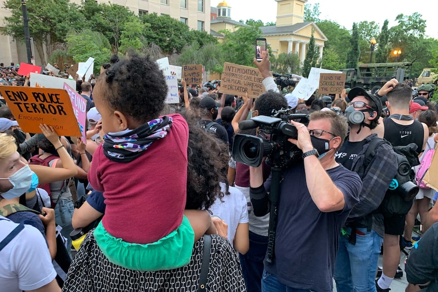 Camera crew wearing masks filming protesters holding up signs and child on shoulders of one person.