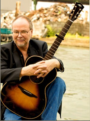 A 2012 photo of Walter Becker of Steely Dan smiling while holding an acoustic guitar.