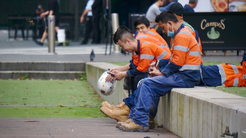 Tradesmen, some wearing masks, sitting and taking a break.