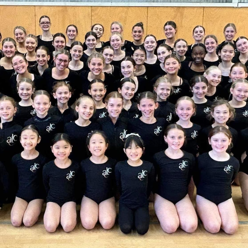 About 50 young girls in black leotards pose in a group smiling in a gumnasium