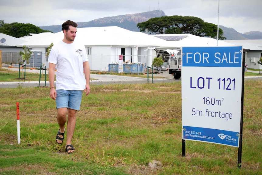 A young man walks towards a for sale sign on a grassy vacant lot.