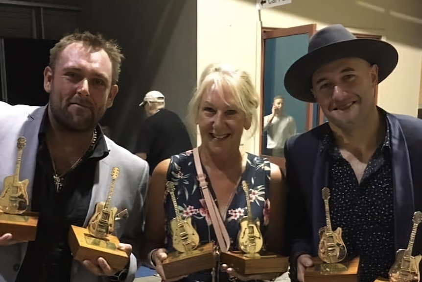 Two men stand with their mother holding golden guitar awards