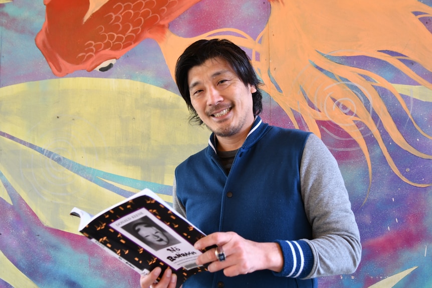 A smiling, bearded man holds a book open while standing in front of a large painting.