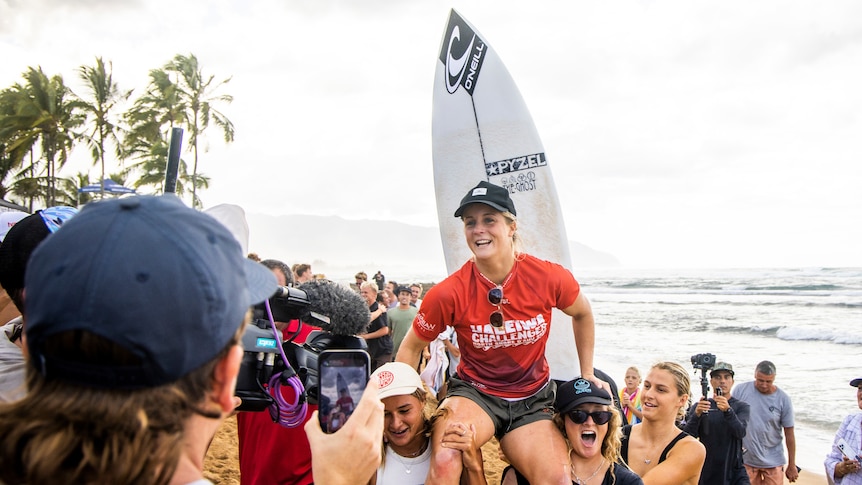 Woman in red smiling, being carried on shoulders, with surfboard behind her